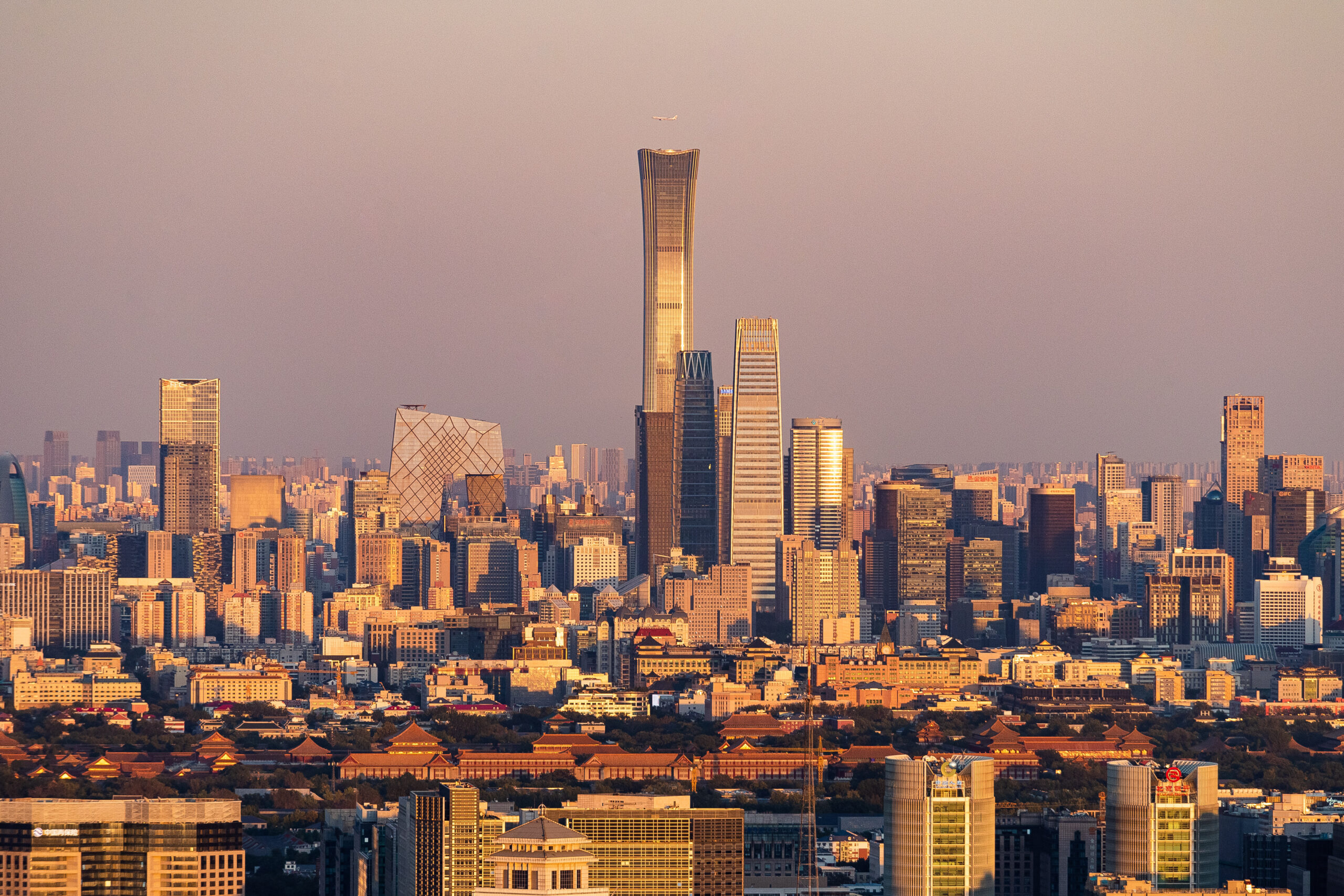 Wide landscape view of Beijing’s Central Business District at sunset, with the tall CITIC Tower (China Zun) centered among skyscrapers and a commercial airliner silhouetted above.